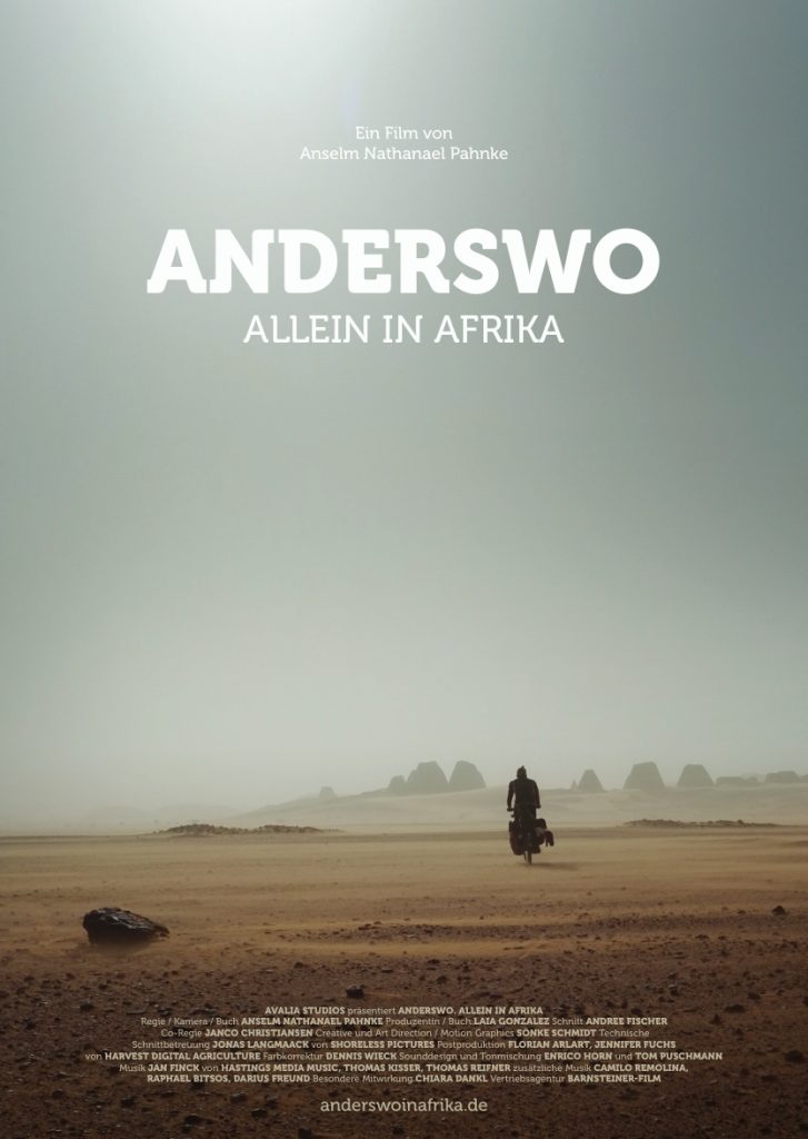 Poster mit dem TItel Anderswo, allein in Afrika. Im Hintergund ist eine weite Wüstenlandschaft zu sehen, davor ein Radfahrer